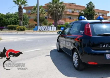 Auto dei carabinieri parcheggiata vicino al cartello di ingresso a Pozzallo, con palme e edifici sullo sfondo