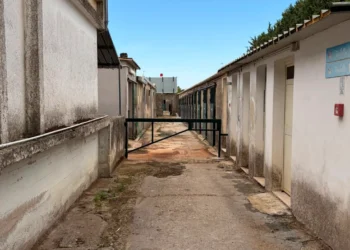 Viale del maneggio comunale tra edifici bassi con porte verdi, una sbarra e cartelli toilette; intonaci ammalorati