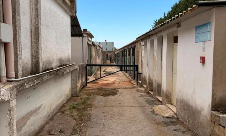 Viale del maneggio comunale tra edifici bassi con porte verdi, una sbarra e cartelli toilette; intonaci ammalorati