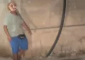 Figura con una maschera dipinta sul volto, maglietta grigia e pantaloncini blu, accanto a un muro con un tubo nero