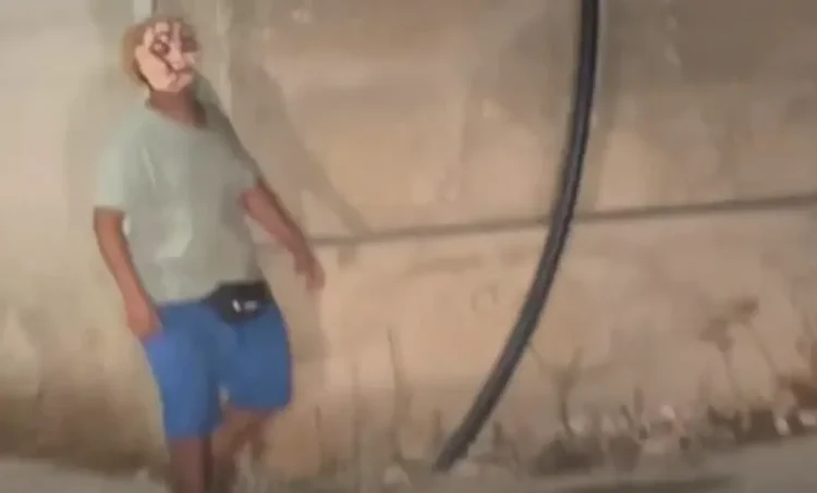 Figura con una maschera dipinta sul volto, maglietta grigia e pantaloncini blu, accanto a un muro con un tubo nero