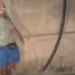 Figura con una maschera dipinta sul volto, maglietta grigia e pantaloncini blu, accanto a un muro con un tubo nero