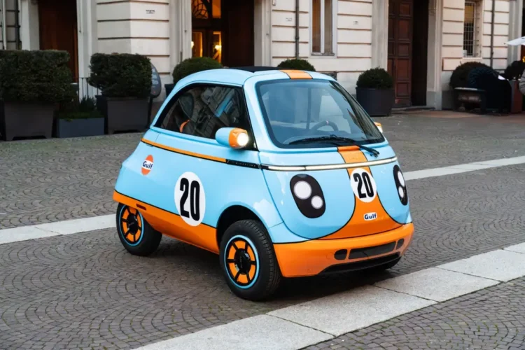 Microlino 777 Collection azzurra e arancione con numero 20 e loghi Gulf, ferma su una strada in pavé davanti a un palazzo