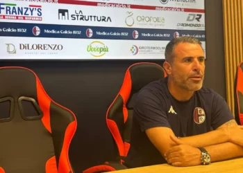 Filippo Raciti, allenatore del Modica, seduto in sala stampa con le braccia sul tavolo, davanti al pannello degli sponsor