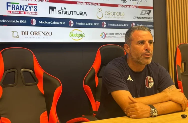 Filippo Raciti, allenatore del Modica, seduto in sala stampa con le braccia sul tavolo, davanti al pannello degli sponsor