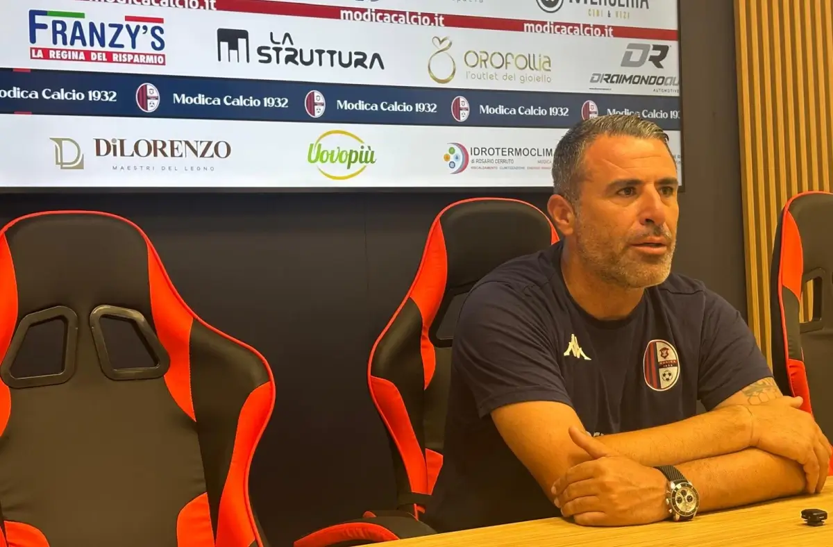 Filippo Raciti, allenatore del Modica, seduto in sala stampa con le braccia sul tavolo, davanti al pannello degli sponsor