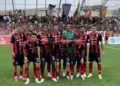 Formazione del Modica schierata in campo prima della gara di Serie D, tribune gremite di tifosi alle spalle