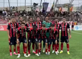 Formazione del Modica schierata in campo prima della gara di Serie D, tribune gremite di tifosi alle spalle