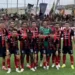 Formazione del Modica schierata in campo prima della gara di Serie D, tribune gremite di tifosi alle spalle