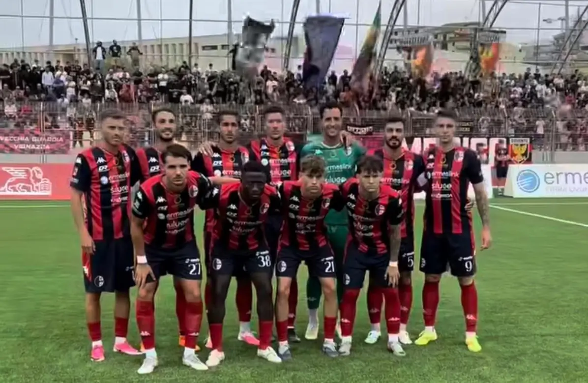 Formazione del Modica schierata in campo prima della gara di Serie D, tribune gremite di tifosi alle spalle