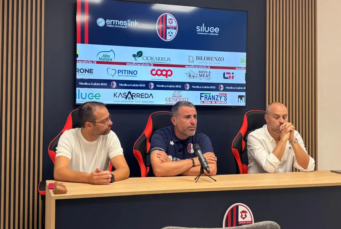 Tavolo della conferenza stampa del Modica Calcio, con logo del club e sponsor sullo schermo alle spalle di tre uomini seduti