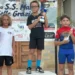 Un giovane ciclista della Motyka Bike School alza il trofeo al centro del podio, tra altri due atleti