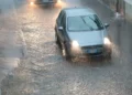 Auto grigia con i fari accesi su una strada allagata sotto un nubifragio; più indietro un'altra vettura con i fari accesi
