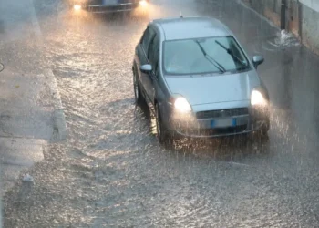 Auto grigia con i fari accesi su una strada allagata sotto un nubifragio; più indietro un'altra vettura con i fari accesi