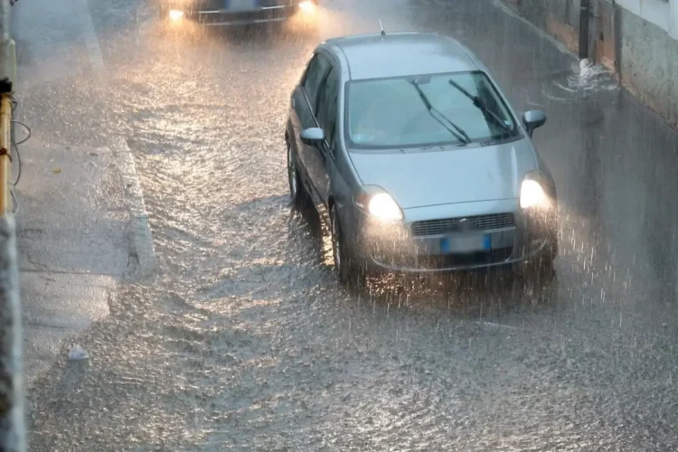Auto grigia con i fari accesi su una strada allagata sotto un nubifragio; più indietro un'altra vettura con i fari accesi