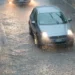 Auto grigia con i fari accesi su una strada allagata sotto un nubifragio; più indietro un'altra vettura con i fari accesi