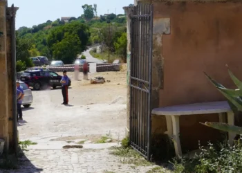 Un carabiniere presidia un cancello con auto dei Carabinieri e persone sfocate dietro il nastro di transennamento