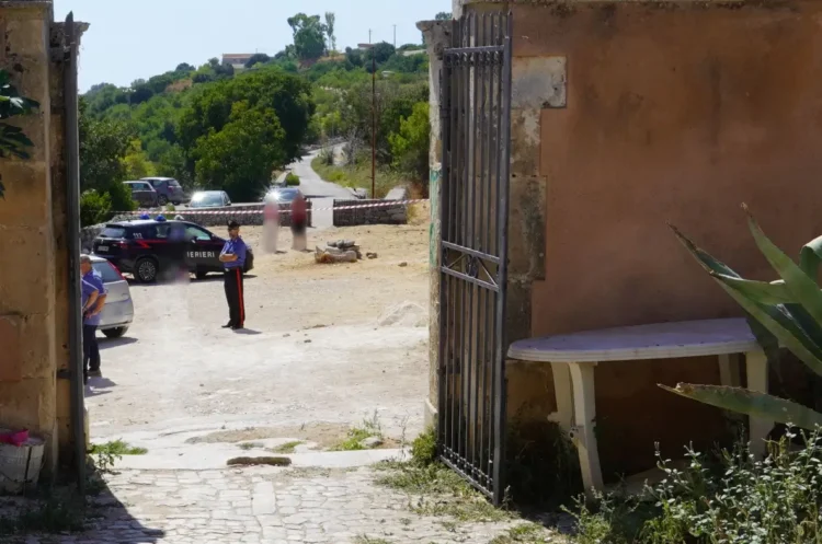 Un carabiniere presidia un cancello con auto dei Carabinieri e persone sfocate dietro il nastro di transennamento