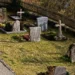 Lapidi e croci di pietra in un cimitero, con steccato in legno sullo sfondo e prato in primo piano