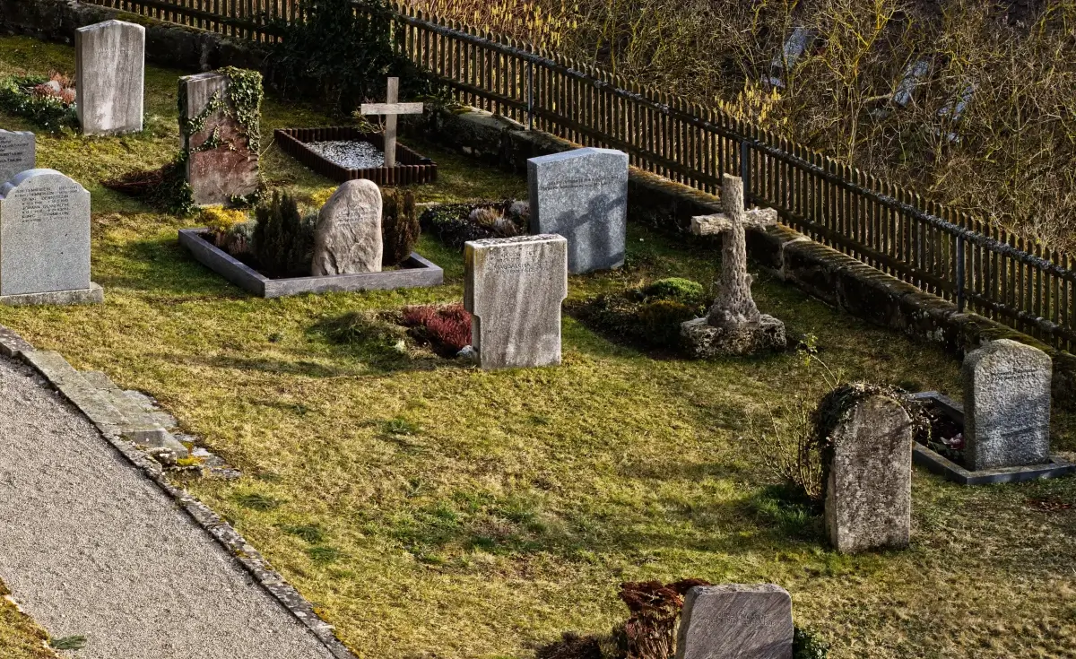 Lapidi e croci di pietra in un cimitero, con steccato in legno sullo sfondo e prato in primo piano