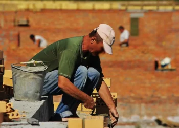 Operaio edile depone mattoni sotto il sole in cantiere, con altri lavoratori sullo sfondo