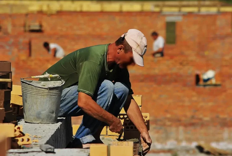 Operaio edile depone mattoni sotto il sole in cantiere, con altri lavoratori sullo sfondo
