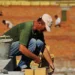 Operaio edile depone mattoni sotto il sole in cantiere, con altri lavoratori sullo sfondo
