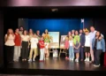 Adulti e bambini in posa sul palco, alcuni con parrucche colorate, accanto a due locandine su cavalletti
