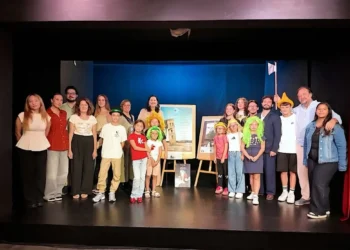 Adulti e bambini in posa sul palco, alcuni con parrucche colorate, accanto a due locandine su cavalletti