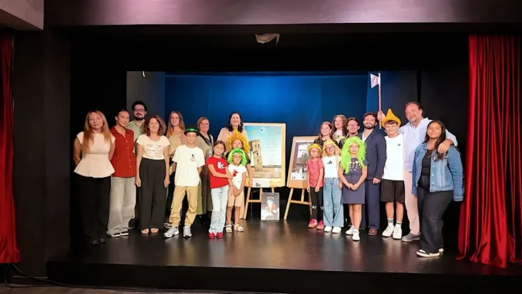 Adulti e bambini in posa sul palco, alcuni con parrucche colorate, accanto a due locandine su cavalletti