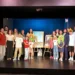 Adulti e bambini in posa sul palco, alcuni con parrucche colorate, accanto a due locandine su cavalletti