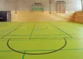Interno di una palestra scolastica con pavimento verde, linee di gioco colorate, porta da calcetto e spalliera a muro