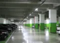 Parcheggio interrato con pilastri bianchi e verdi, auto in sosta ai lati e corsia centrale illuminata da neon