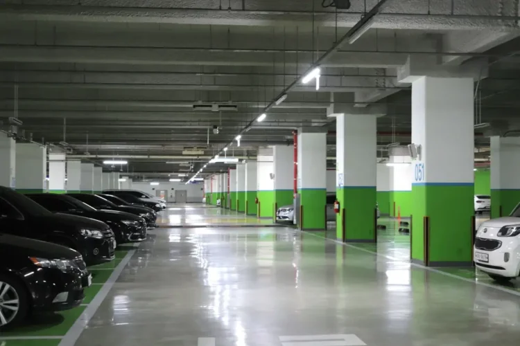 Parcheggio interrato con pilastri bianchi e verdi, auto in sosta ai lati e corsia centrale illuminata da neon