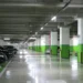 Parcheggio interrato con pilastri bianchi e verdi, auto in sosta ai lati e corsia centrale illuminata da neon