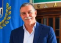Peppe Privitera sorridente, in giacca blu e camicia azzurra, accanto a un gonfalone con rami di quercia