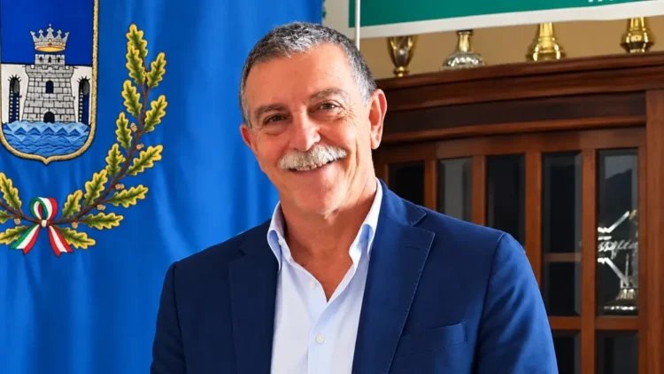 Peppe Privitera sorridente, in giacca blu e camicia azzurra, accanto a un gonfalone con rami di quercia