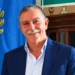 Peppe Privitera sorridente, in giacca blu e camicia azzurra, accanto a un gonfalone con rami di quercia