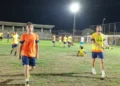 Calciatori con maglie arancioni e pettorine gialle su un campo in erba illuminato di sera, con una tribuna sullo sfondo