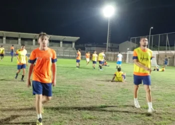 Calciatori con maglie arancioni e pettorine gialle su un campo in erba illuminato di sera, con una tribuna sullo sfondo