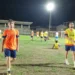 Calciatori con maglie arancioni e pettorine gialle su un campo in erba illuminato di sera, con una tribuna sullo sfondo