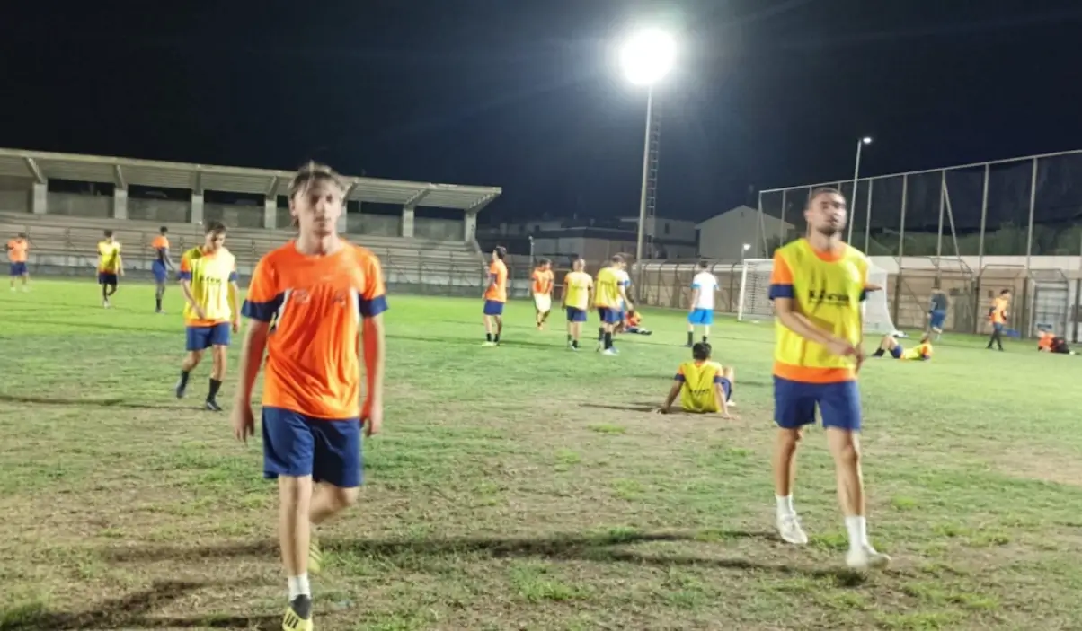 Calciatori con maglie arancioni e pettorine gialle su un campo in erba illuminato di sera, con una tribuna sullo sfondo