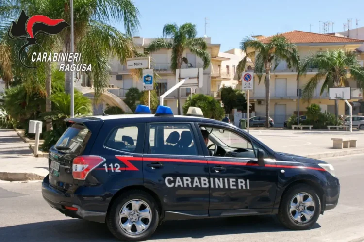 Auto dei carabinieri di Ragusa in transito in una piazza con palme, tra abitazioni e cartelli stradali