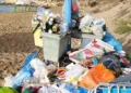 Bidoni stracolmi e cumuli di rifiuti abbandonati sulla sabbia di una spiaggia, con sdraio e ombrelloni sullo sfondo