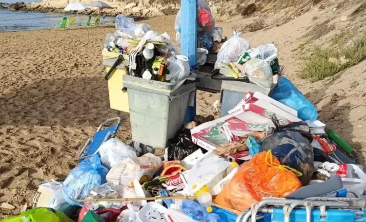 Bidoni stracolmi e cumuli di rifiuti abbandonati sulla sabbia di una spiaggia, con sdraio e ombrelloni sullo sfondo