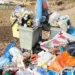 Bidoni stracolmi e cumuli di rifiuti abbandonati sulla sabbia di una spiaggia, con sdraio e ombrelloni sullo sfondo