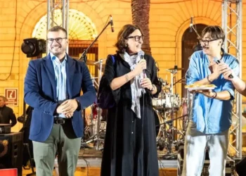 Sul palco di Scacciapensieri a Modica, tre persone con il microfono; una tiene in mano un piatto con una scaccia