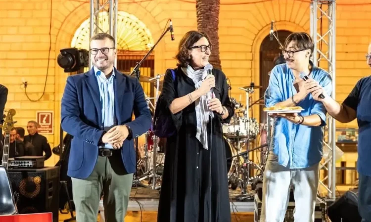 Sul palco di Scacciapensieri a Modica, tre persone con il microfono; una tiene in mano un piatto con una scaccia