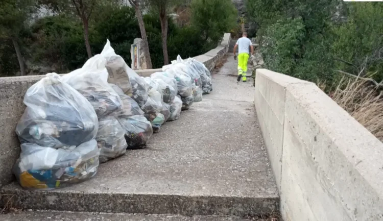 Sacchi di rifiuti allineati sul muretto della scala di San Francesco a Ispica, con un operatore ecologico che sale i gradini
