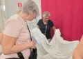 Due donne osservano un ampio tessuto bianco lavorato a sfilato, aperto davanti a un telo rosso, in una sala con vetrine